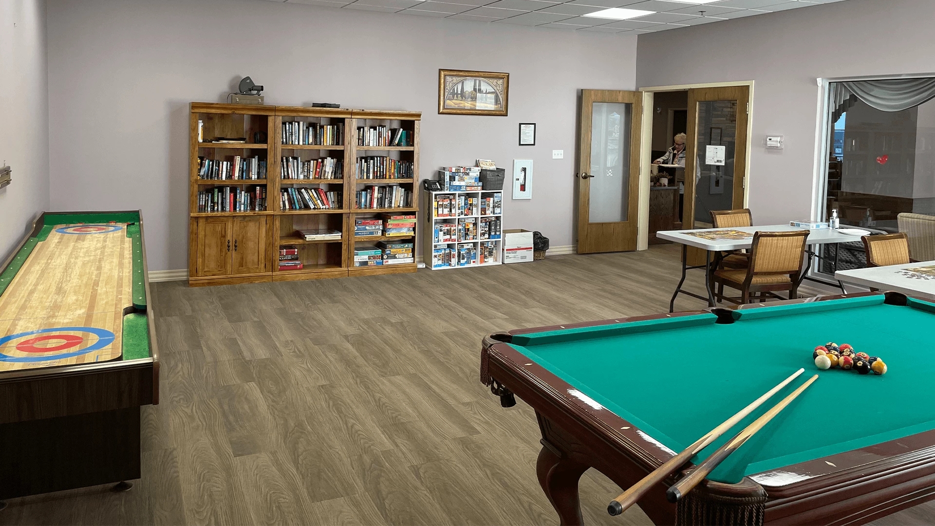 Games room at Cedarwood. There is a shelf with books, a shelf with games, shuffleboard, a pool table, and a table with puzzles on it.